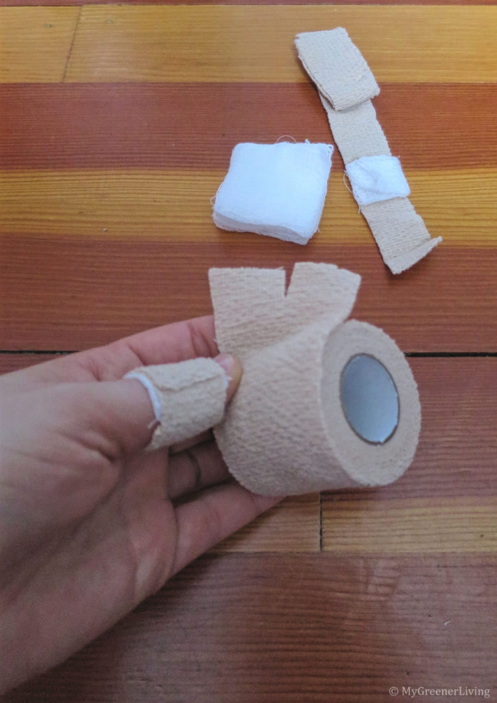 self-adhesive bandage cut into 1" wide strips for first aid bandages, with bandage wrapped around thumb and another strip of self-adhesive bandage with gauze in the background