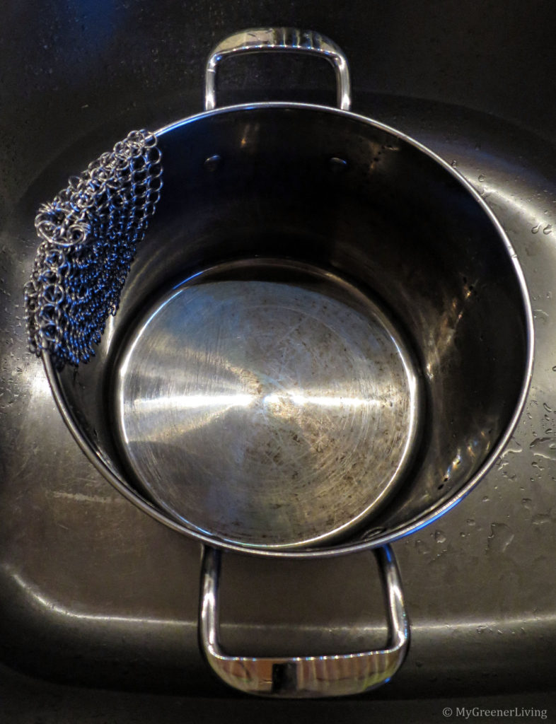 Photo of a pot that has had the left half scrubbed with a stainless steel chain mail scrubber, soap, and water, and the right half just cleaned with a regular kitchen brush, soap, and water. The left half is shiny silver and the left half had brownish yellow stains. The chain mail scrubber is hanging over the edge of the pot.