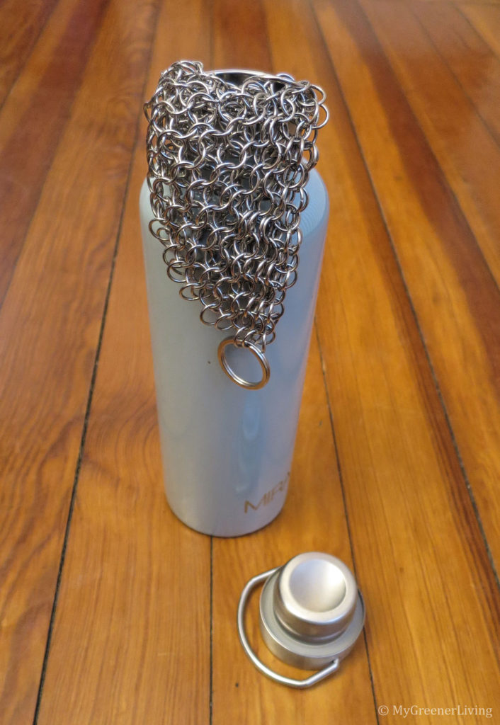 Photo of a stainless steel reusable water bottle with stainless steel chain mail scrubber hanging half in, half out of the bottle.